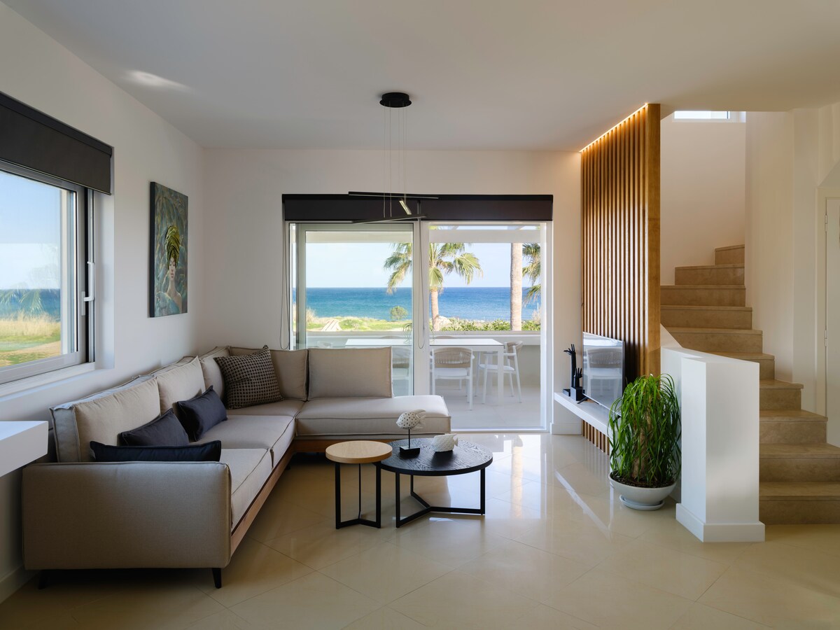 A modern living area features a spacious sectional sofa with neutral-toned cushions. A coffee table is placed in front, and large windows offer unobstructed views of the sea and surrounding greenery. Soft natural light illuminates the space, complemented by a wooden staircase leading to the upper level.