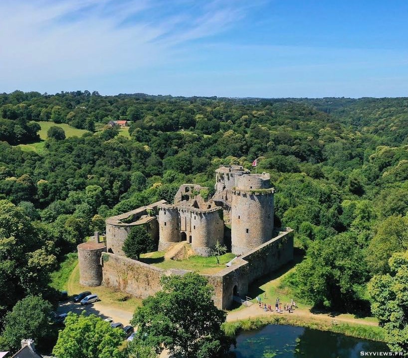 A historic castle is set against a backdrop of lush greenery and blue skies. The structure features multiple towers and fortified walls, surrounded by trees and a serene water body. A glimpse of parking is visible in the foreground, enhancing accessibility to this scenic location.