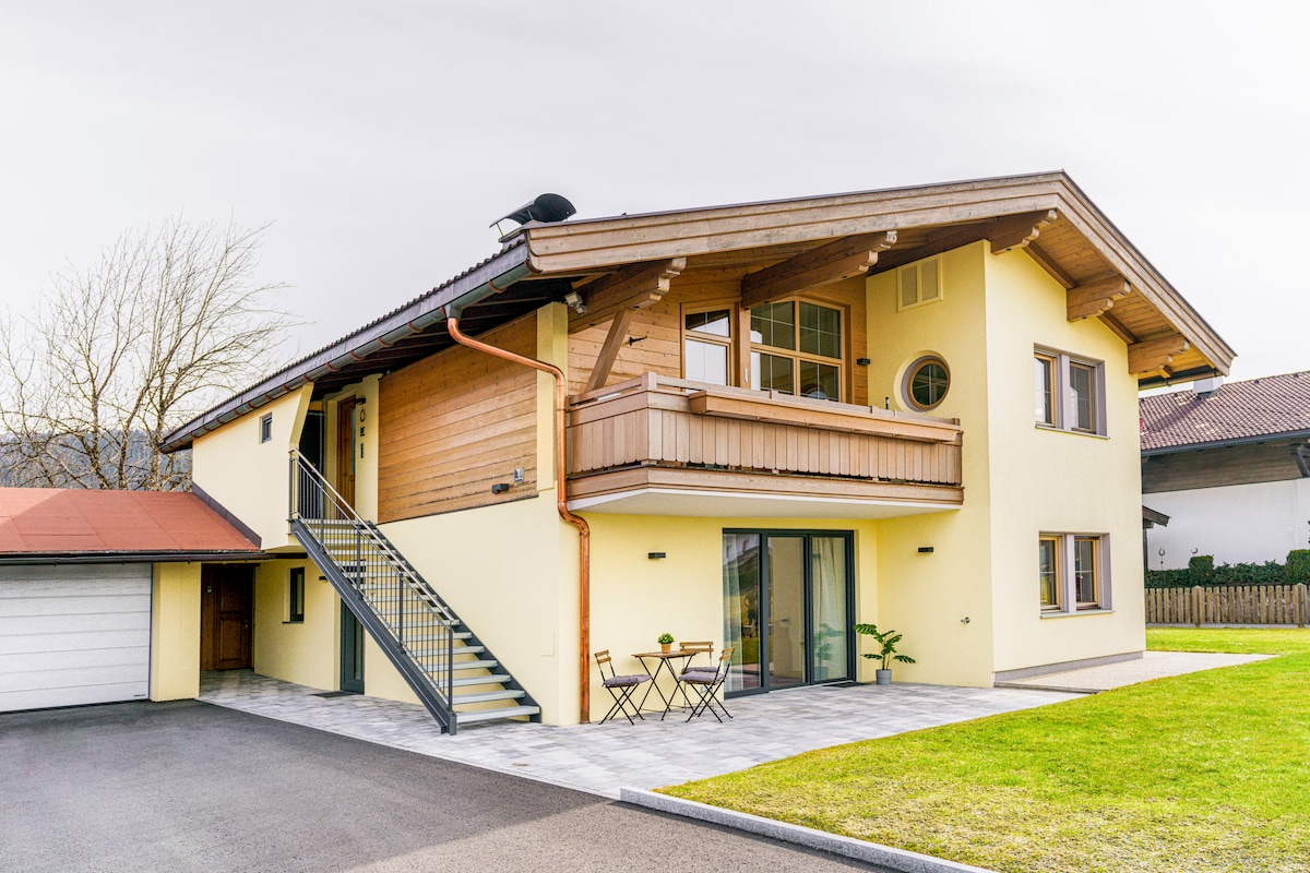 The two-story house features a combination of yellow and wooden exterior elements. A balcony extends from the upper level, while a set of stairs leads to the entrance. The driveway is paved, bordered by a well-maintained lawn and small outdoor seating area.