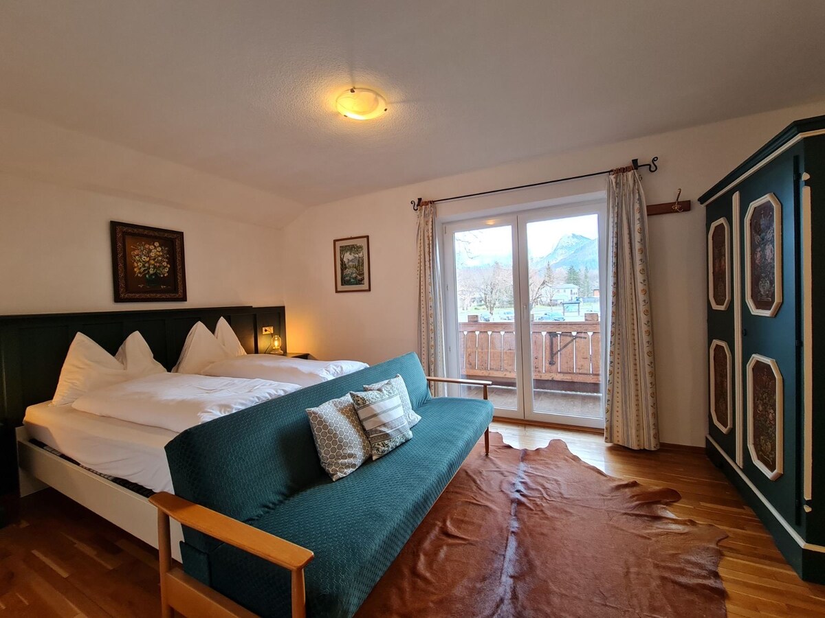 A cozy room features a spacious bed with plush white bedding and two decorative pillows. A blue sofa is positioned in front of large glass doors leading to a balcony. Natural wood flooring complements the simple furnishings, while artwork adorns the walls.