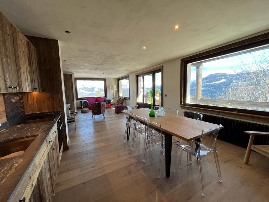 The dining area features a long wooden table surrounded by clear chairs. Large windows offer expansive views of the mountains. A cozy seating arrangement with comfortable chairs is visible in one corner, while natural light fills the space, enhancing the warm wooden tones.