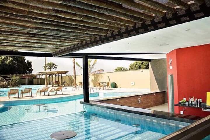 An indoor area features a sleek bar with a pool view, surrounded by generous glass walls. Sunlight brightens the space and highlights the blue, tiled swimming pool beyond, where lounge chairs are positioned under a sheltered area, enhancing the inviting ambiance.