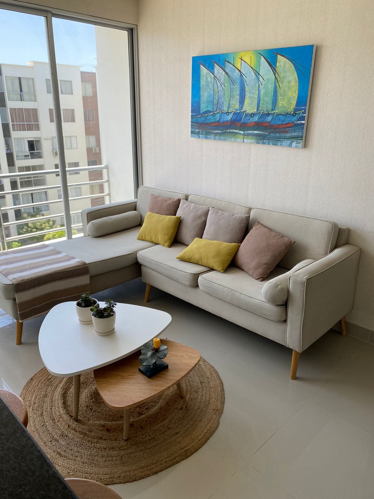 A light-filled living area features a soft, beige sofa adorned with colorful cushions. A round, woven rug is situated beneath a modern coffee table, which holds small decorative items. A vibrant painting of sailboats adds a touch of color to the wall, while a view of the outdoors is visible through large windows.