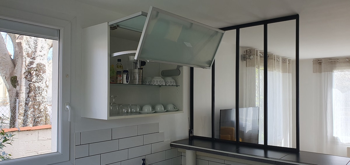 A modern kitchen area is visible, featuring a large window that allows natural light to fill the space. The cabinetry includes a lift-up section, revealing a selection of glassware and storage space. A sleek black divider separates the kitchen from the living area.