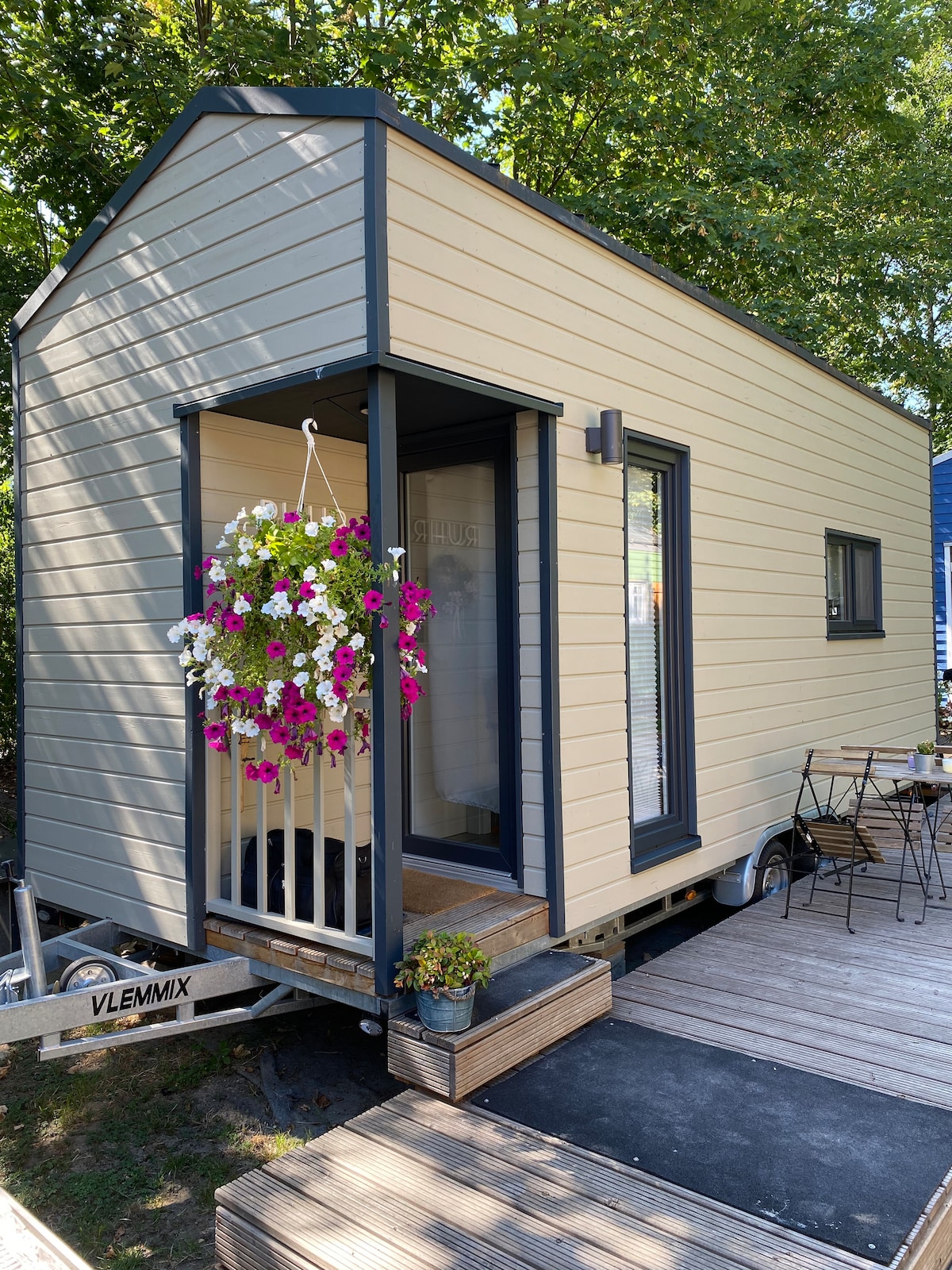 A modern tiny house with a light-colored exterior is framed by greenery. A welcoming entrance features a hanging flower basket filled with vibrant blooms. A small outdoor seating area with a table and chairs is positioned on the wooden deck beside the house.