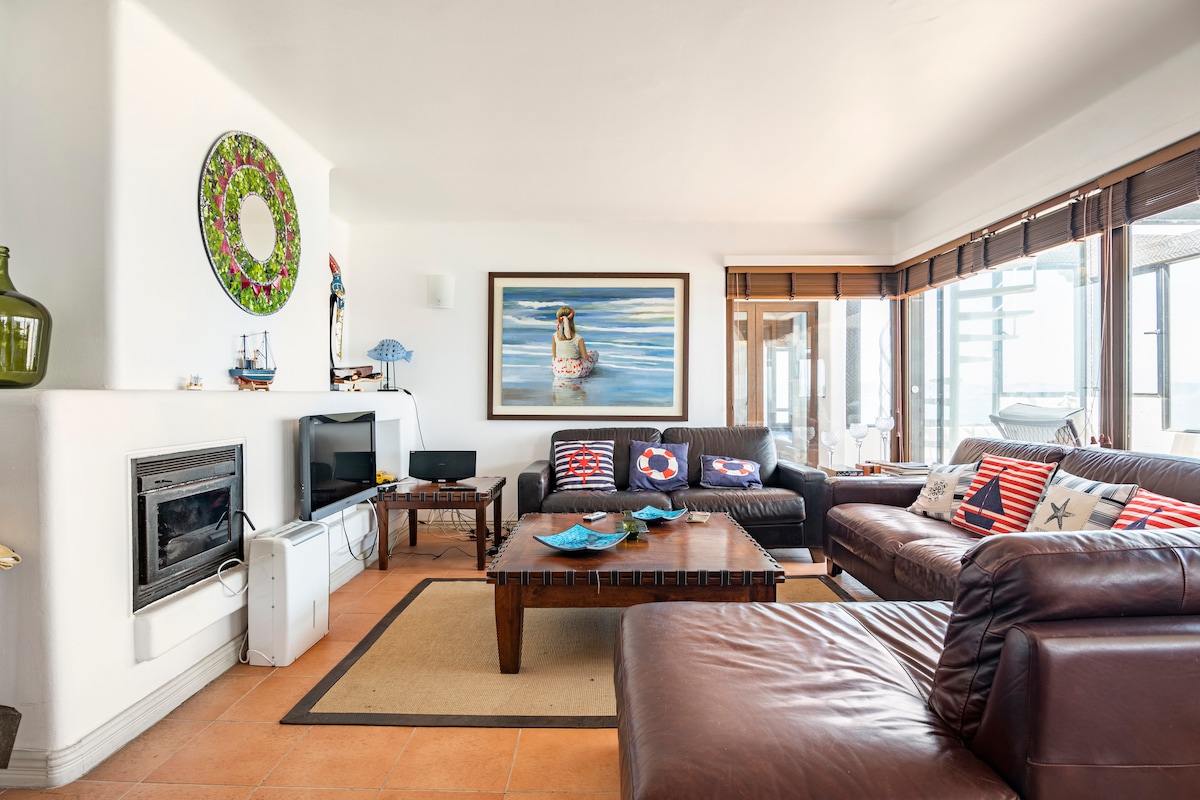 A spacious living area features a mix of comfortable seating arrangements with earth-toned leather sofas and decorative throw pillows. A wooden coffee table is positioned centrally, and art adorns the walls. Natural light filters through large windows, enhancing the welcoming ambiance.
