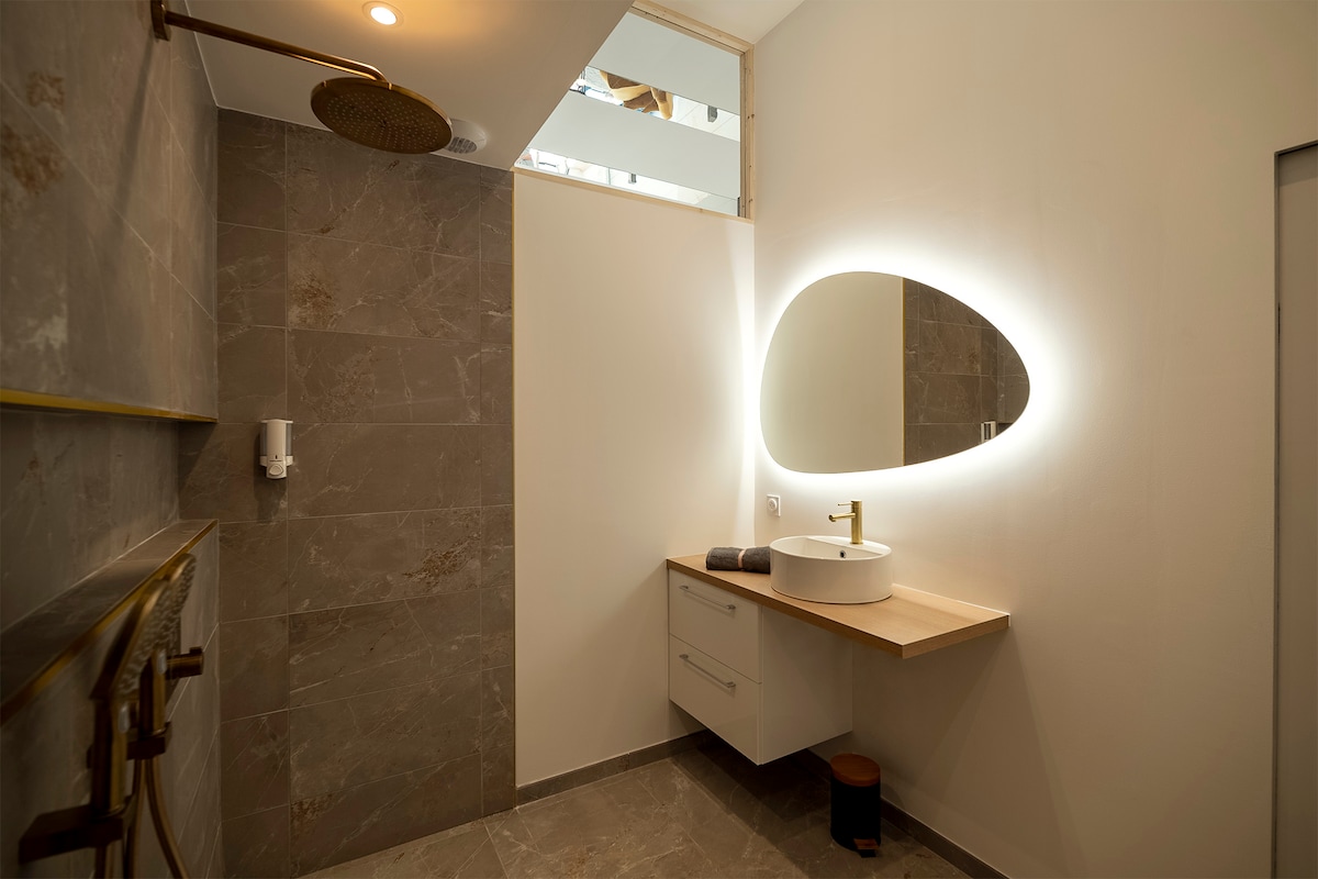 A modern bathroom features elegant gray stone walls and a spacious layout. A sleek oval mirror, illuminated by soft lighting, complements the stylish sink with a gold faucet. A minimalist countertop and a wooden stool add functional elements, enhancing the overall contemporary design.