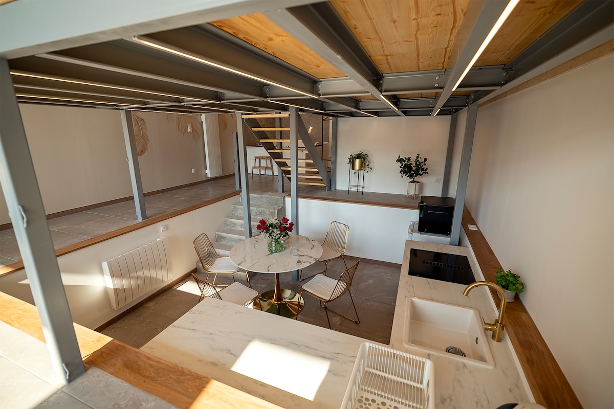 An open-plan kitchen and dining area is displayed, featuring a round marble table surrounded by four gold metal chairs. Above, a staircase leads to a lofted space, complemented by a minimalist décor with a potted plant and subtle lighting enhancing the inviting atmosphere.