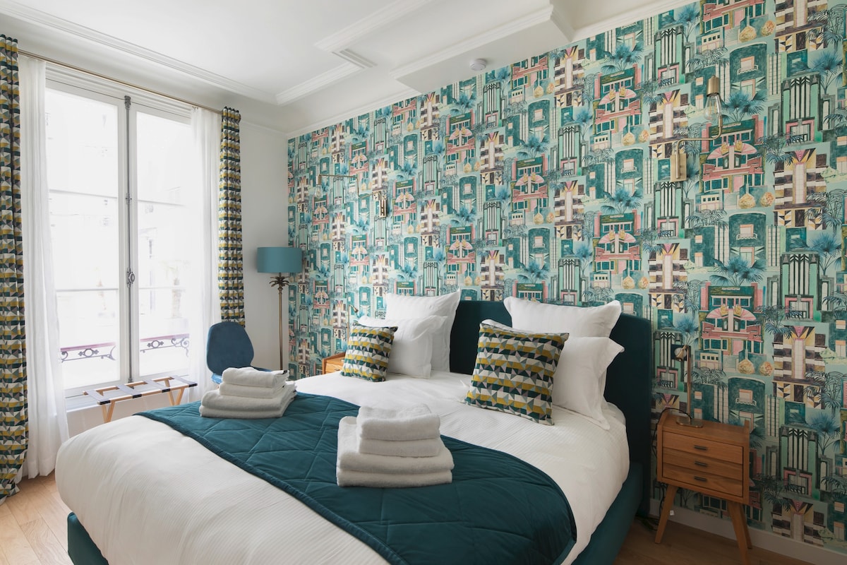 The bedroom features a spacious bed dressed in white linens and accented by colorful throw pillows. A vibrant, patterned wallpaper complements the decor. Natural light floods in through large windows, framed with curtains. A small bedside table and chair complete the stylish setting.
