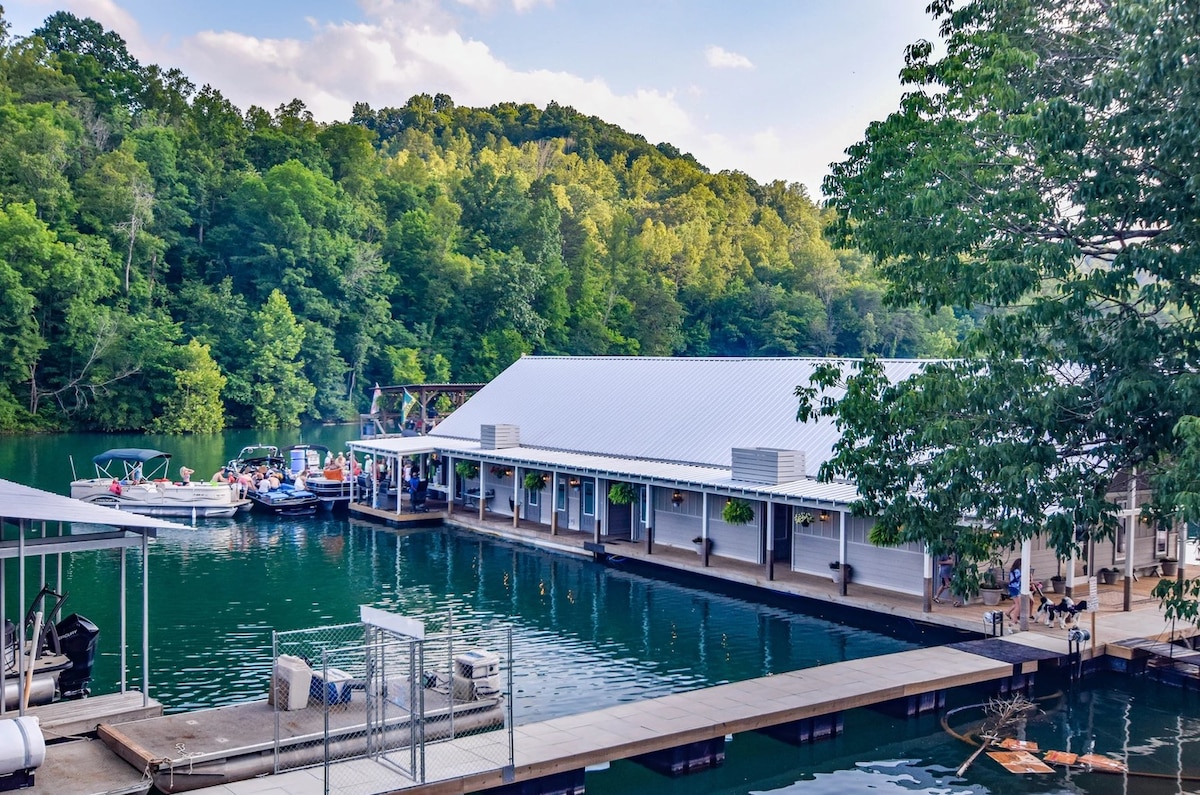 A floating home is shown, nestled in a serene marina with lush green hills in the background. The structure features a modern roof and large windows. Nearby boats are docked, and a wooden walkway leads to the entrance, enhancing the connection to the water.
