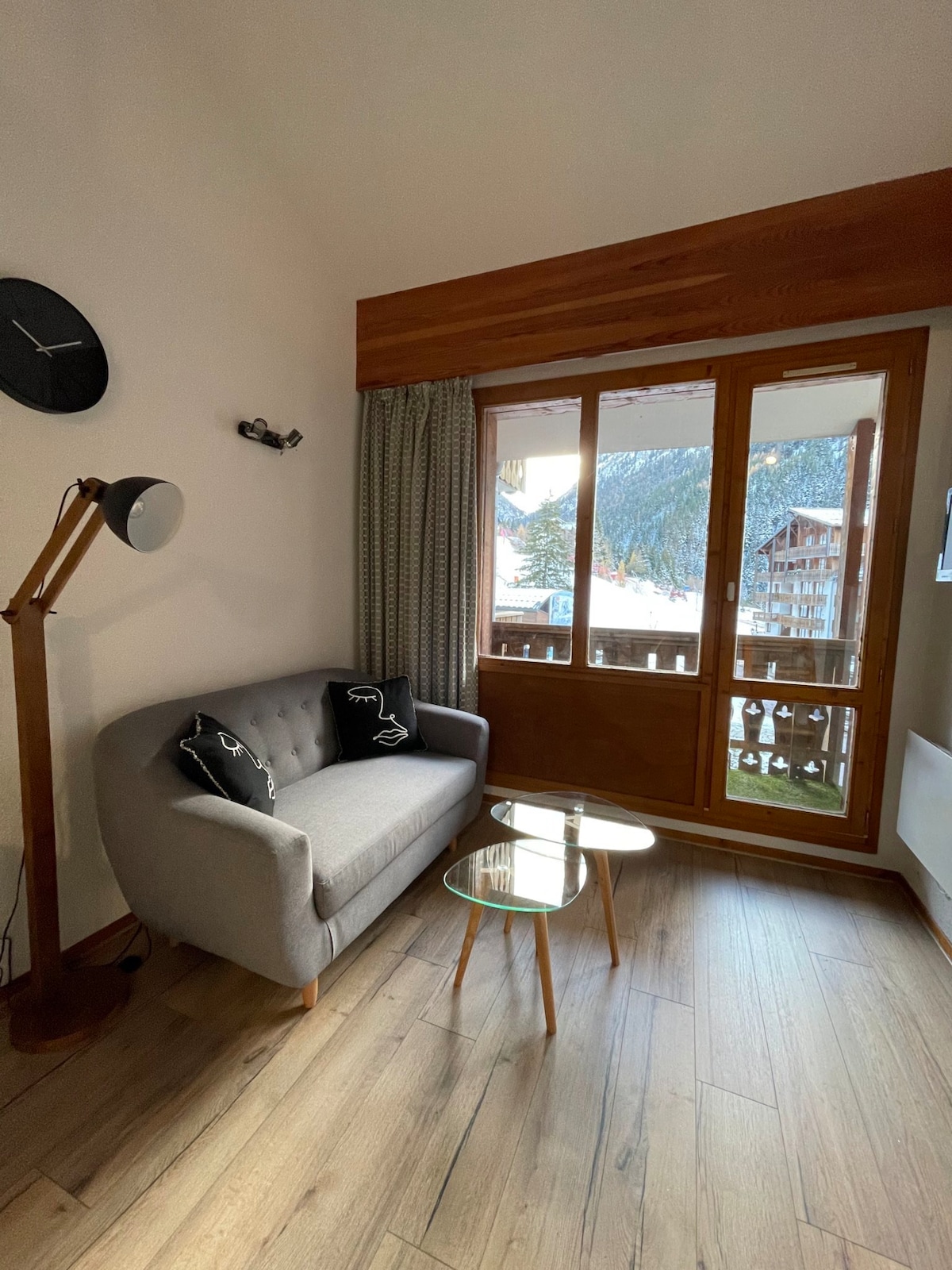 A cozy living area features a grey upholstered sofa paired with two small glass tables. A wall clock is visible above the sofa, and the large windows offer a view of the exterior landscape, allowing natural light to fill the space.