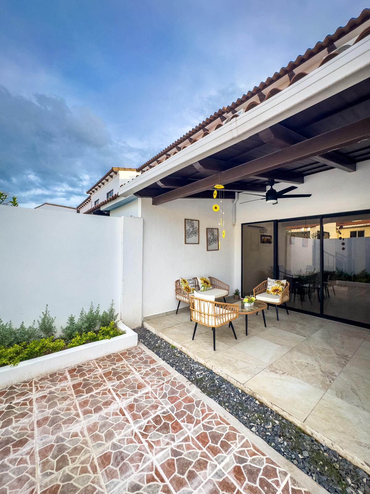 A charming outdoor seating area features a set of light-colored wicker chairs complemented by a small table adorned with fresh greenery. The space is framed by a stone path and decorative plants, with a glimpse of the house's architecture visible in the background.