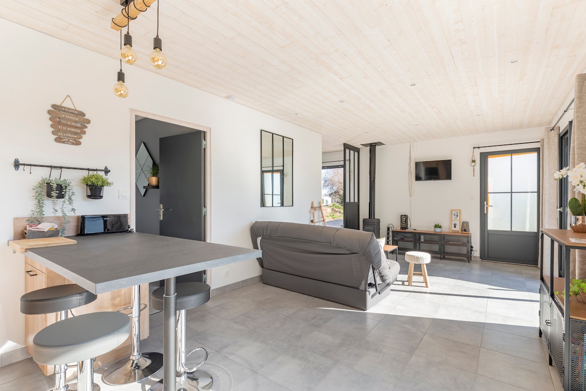 A bright and airy open-plan living space features a cozy seating area with a grey sofa and a minimalist coffee table. A dining area with modern stools is situated beside a well-equipped kitchen, illuminated by natural light through large windows. A door leads to the outdoor area.