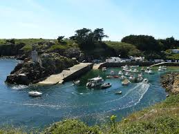 A calm harbor scene is depicted with several boats anchored in the water. The shoreline is lined with rocky outcrops and greenery. A small pier extends into the water, and a lighthouse is visible against a clear blue sky.