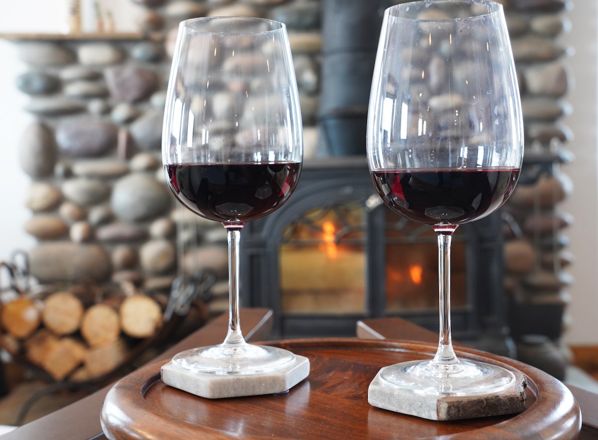 Two elegant wine glasses filled with red wine are positioned on a wooden tray. A stone fireplace in the background is softly illuminated, casting a warm glow. Stacked logs are visible beside the fireplace, creating a cozy setting.