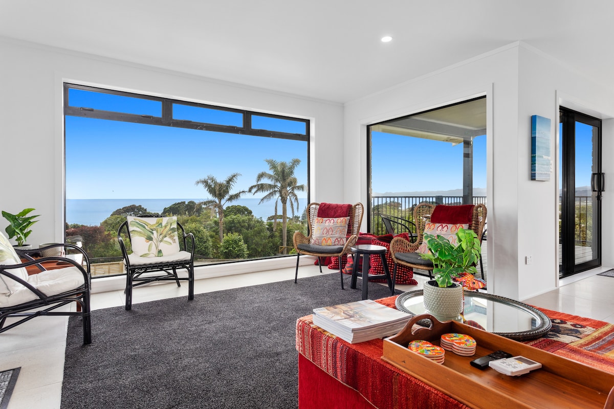 A bright and airy living area features large windows offering panoramic views of the sea and surrounding greenery. Two comfortable armchairs and a stylish couch provide seating, while a low coffee table holds decorative items. A potted plant adds a splash of greenery to the space.
