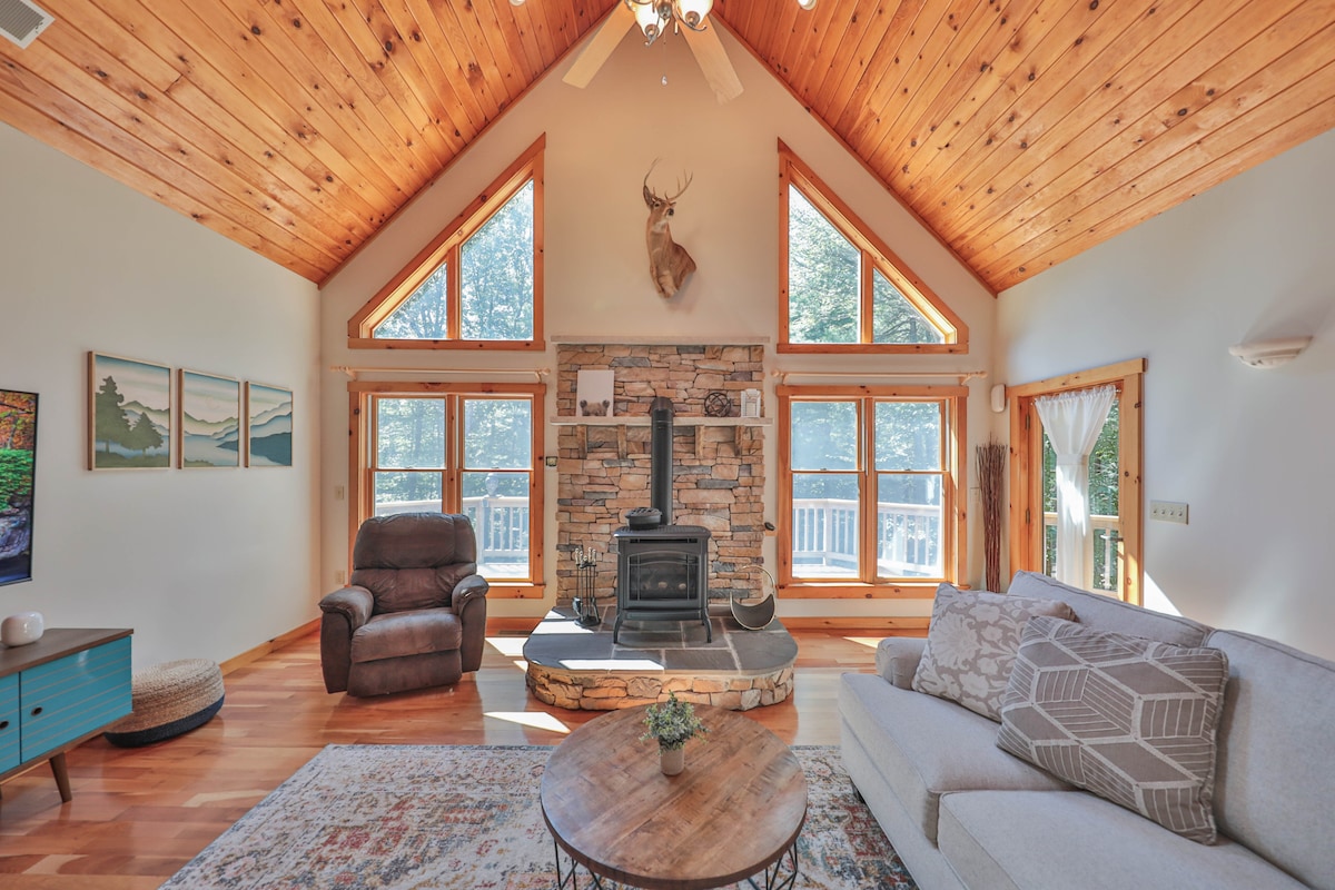 A spacious living area features a wooden vaulted ceiling with large triangular windows allowing natural light. A central stone fireplace adds warmth, and comfortable seating is positioned around a circular coffee table. Scenic outdoor views are framed by the windows, creating a calm ambiance.