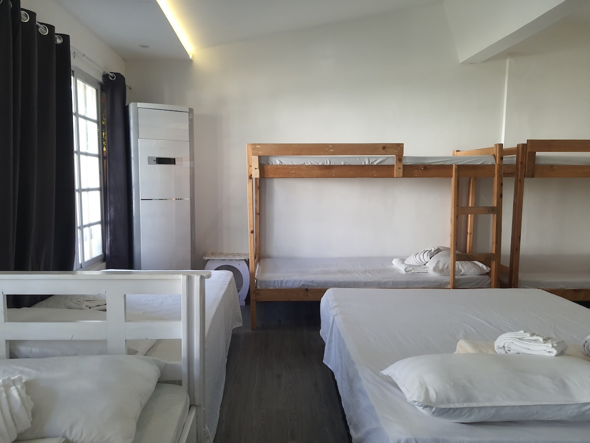 The room features a mix of bunk beds and a spacious bed, creating ample sleeping options. Natural light fills the room through large windows with dark curtains. Neat bedding is arranged on each bed, maintaining a tidy appearance.