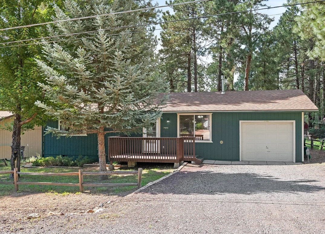 The front exterior of a charming cabin is shown, featuring a green facade and a brown shingled roof. A small deck with a railing is visible, leading to the main entrance. Surrounding trees add a natural element to the backdrop, while a gravel area and a single-car garage are present.