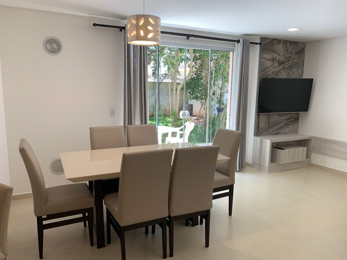 A dining area features a light-colored table surrounded by six beige chairs. Natural light enters through large sliding glass doors, which open to a garden area. A flat-screen TV is mounted on the wall, and a comfortable entertainment unit is positioned below.