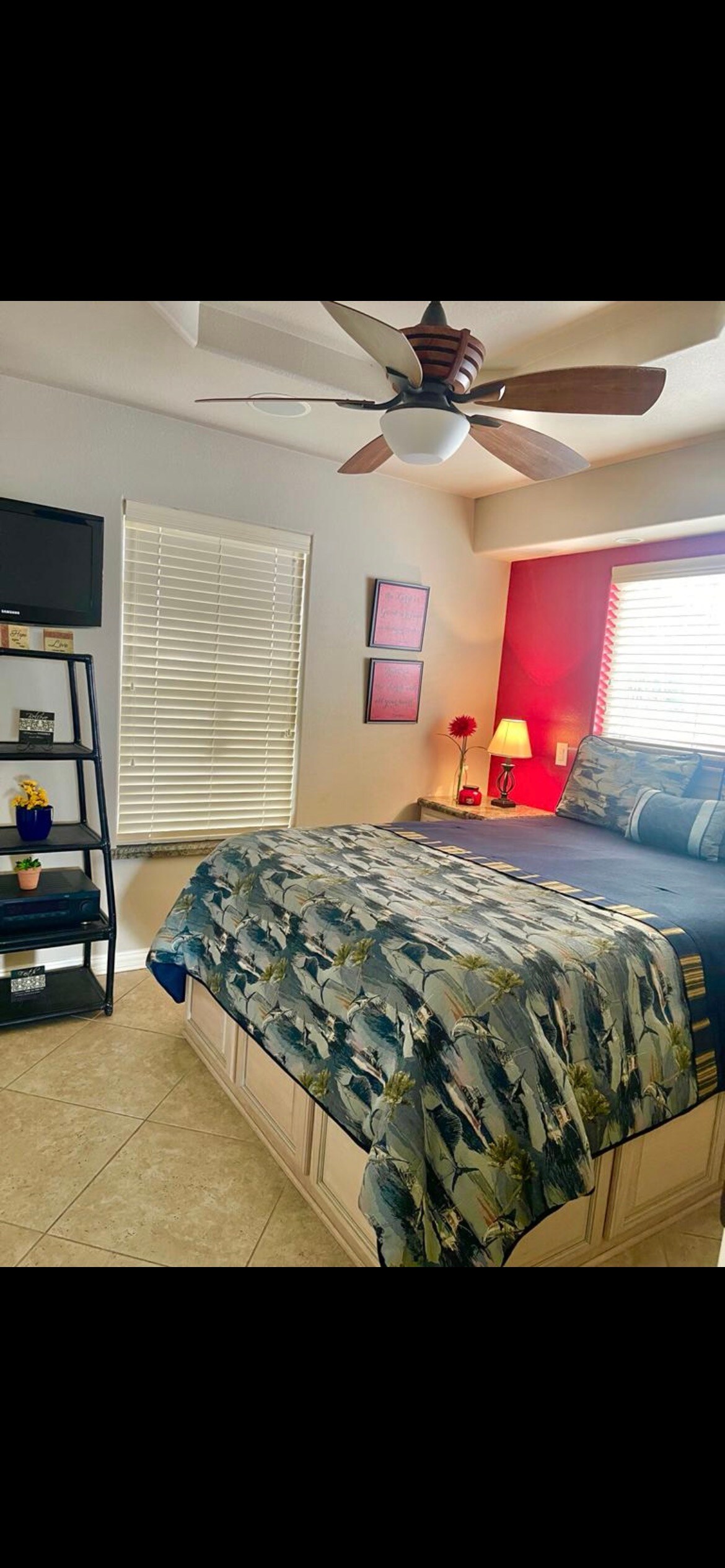 A cozy bedroom featuring a spacious bed with a patterned blue and green quilt. A ceiling fan is mounted overhead, and natural light filters through white blinds. A small TV is positioned on a black shelving unit, accompanied by decorative items and a lamp.