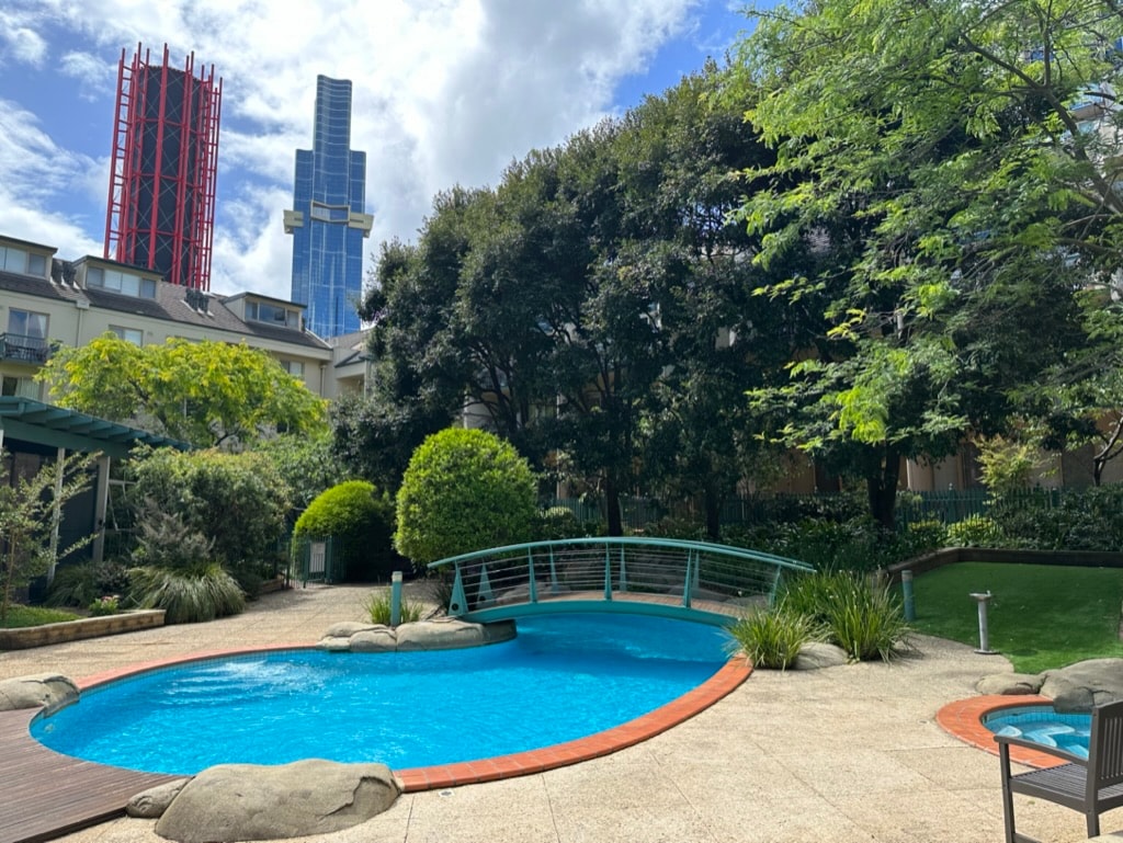 A landscaped outdoor pool area features a curved blue swimming pool surrounded by greenery. Smooth stones frame the pool, while a wooden bridge leads to a grassy area. Tall buildings are visible in the background under a partly cloudy sky.