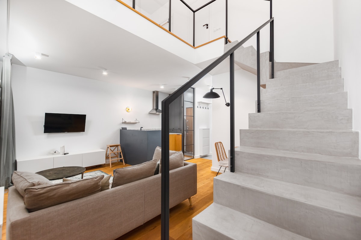 A spacious living area is presented with a comfortable sofa and a flat-screen TV mounted on the wall. Stairs with a sleek design lead to the upper level, while the kitchen is visible in the background, featuring modern appliances and a minimalist aesthetic.