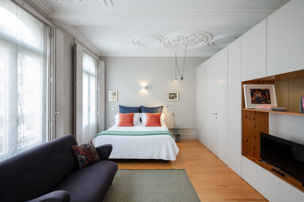 A spacious studio features a comfortable bed with decorative pillows, complemented by a soft green blanket. Natural light filters through large windows, highlighting elegant architectural details. A cozy seating area and a stylish entertainment unit create a harmonious living space.