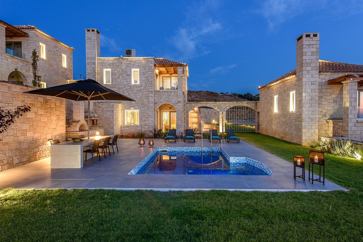 An outdoor area features a stone patio with a swimming pool, surrounded by comfortable lounge chairs and an umbrella-shaded dining table. The villas are seen in the background, constructed with a natural stone facade, and well-maintained landscaping enhances the inviting atmosphere.