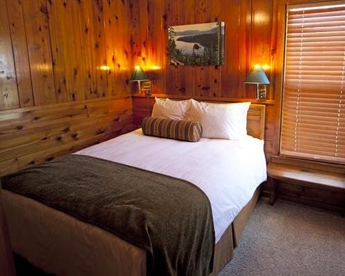 A cozy bedroom features warm wooden paneling, providing a natural ambiance. A spacious bed with white linens is centered in the room, flanked by two bedside lamps. A scenic landscape picture adorns the wall above the headboard, and a small bench sits beside a window with closed blinds.
