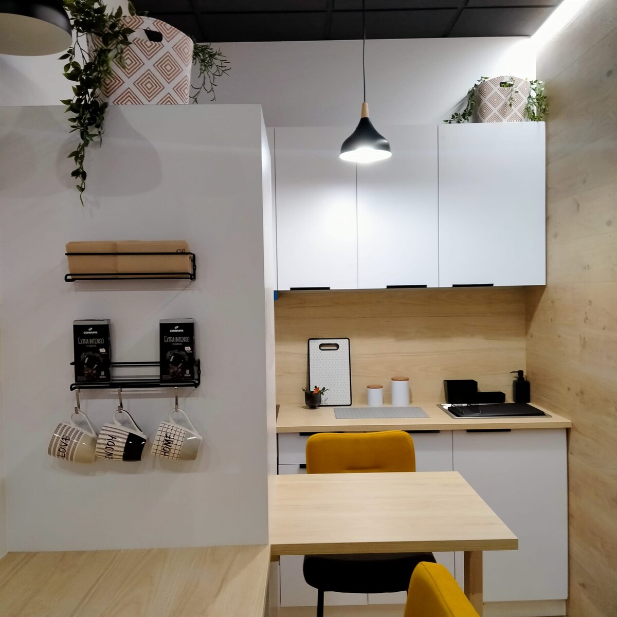 A modern kitchen area is presented, featuring sleek white cabinetry and wooden accents. A small table with a yellow chair complements the space. Cooking essentials are organized neatly, with dishware and utensils hanging above the countertop, illuminated by a pendant light overhead.