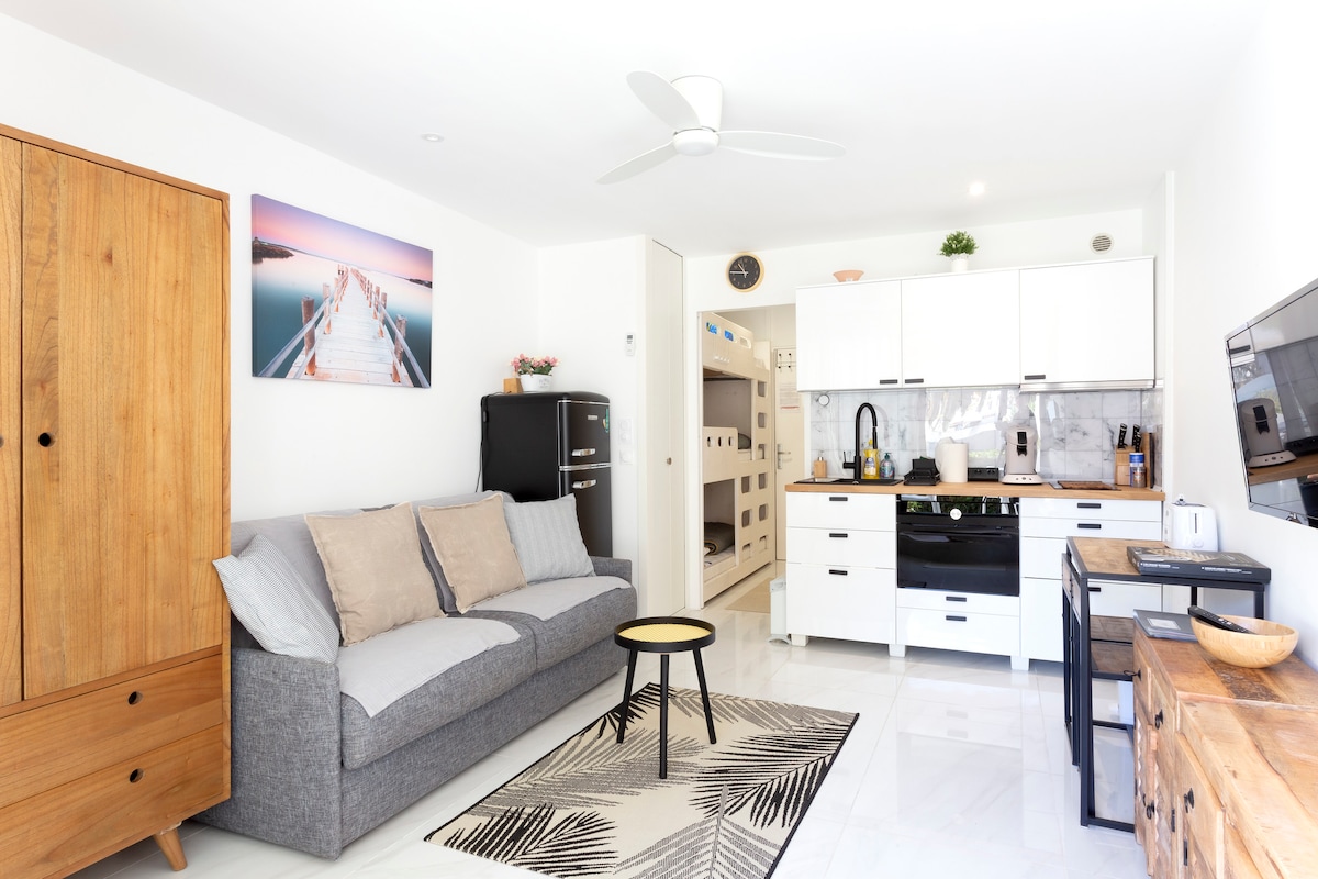 A light-filled living area features a gray sofa adorned with decorative pillows. A round coffee table sits on a patterned rug, while a wall-mounted TV and a small black refrigerator are visible. The open kitchen includes modern appliances and a stylish backsplash.