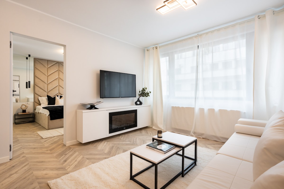 A bright sitting area features a light sofa positioned near a sleek coffee table. A large television is mounted on the wall, and ample natural light streams in through expansive windows with sheer curtains. The space is connected to a room visible in the background.