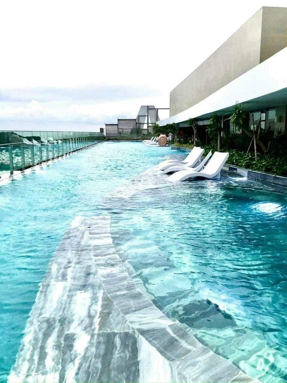 An infinity pool is featured at the rooftop, with clear blue water extending towards the horizon. Lounge chairs are positioned along the poolside, complementing the modern design. Lush greenery is visible along the surrounding edges, creating a serene environment.