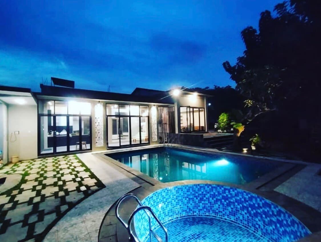 An evening view showcases a modern villa with large glass doors opening to an inviting pool area. The pool is illuminated, reflecting the evening sky. Surrounding greenery contributes to a serene atmosphere.