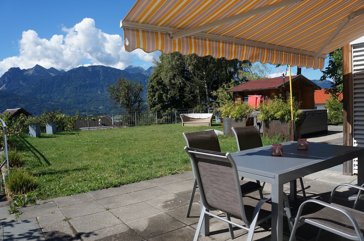 An outdoor terrace features a dining table with chairs, shaded by a striped awning. The surrounding area includes a grassy lawn and a view of distant mountains under a partly cloudy sky.