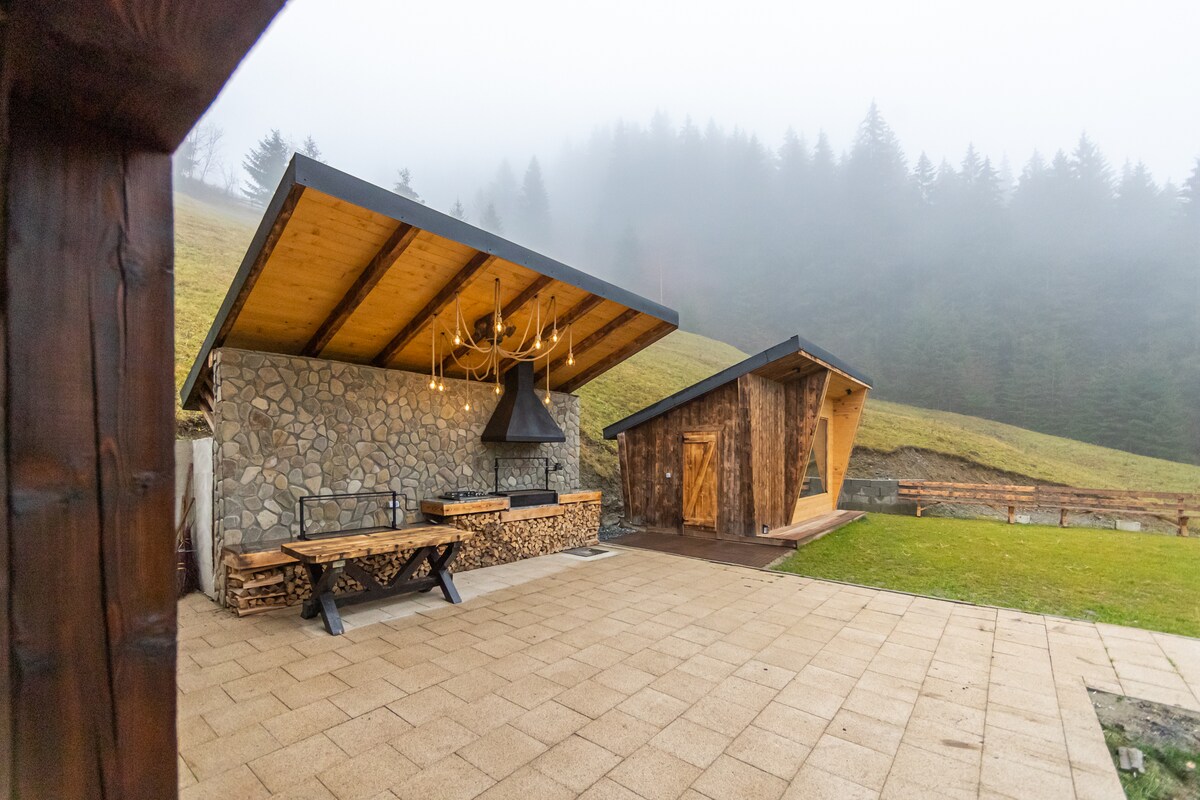 An outdoor area features a stone grill station with a wooden table and bench, surrounded by a spacious patio. A wooden cabin sits nearby under a sloping roof, with misty mountains in the background, creating a serene atmosphere.
