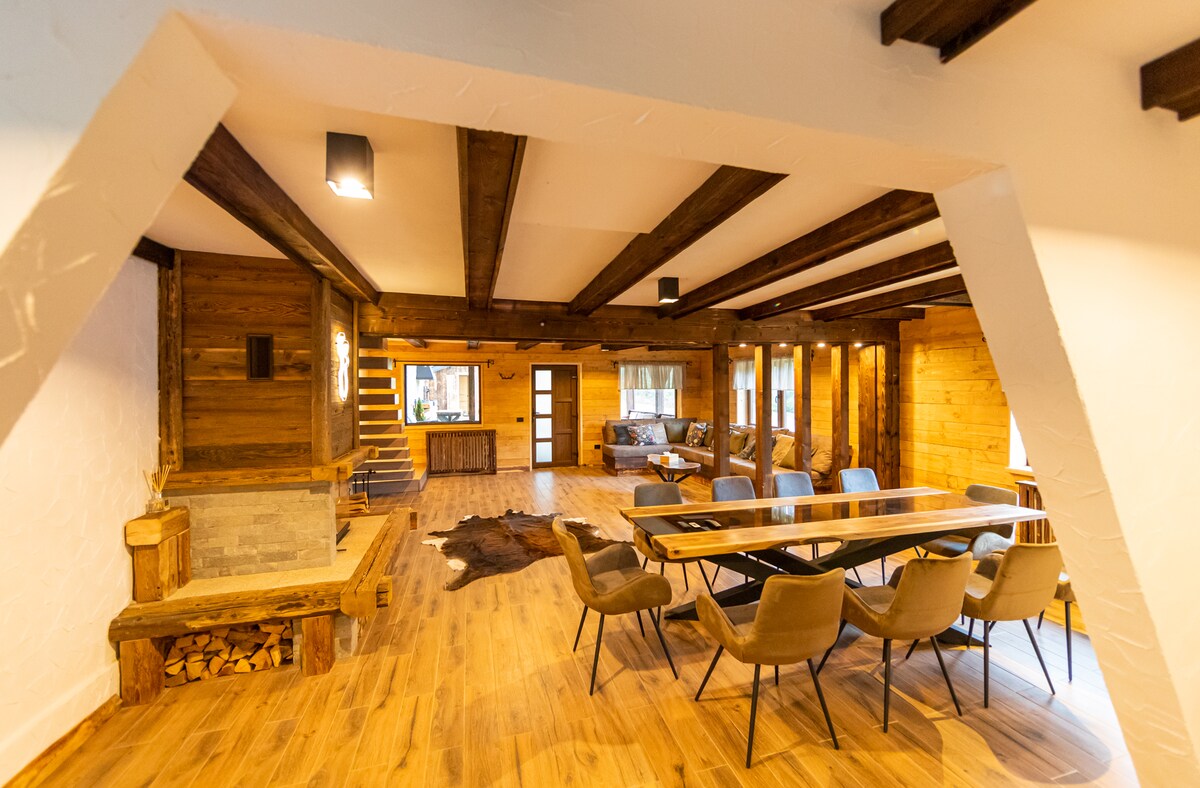 A spacious living area features wooden beams and a combination of rustic and modern design elements. A large dining table accompanied by plush chairs is positioned near a cozy seating area. Natural light floods the room, highlighting the wood finishes and warm tones.