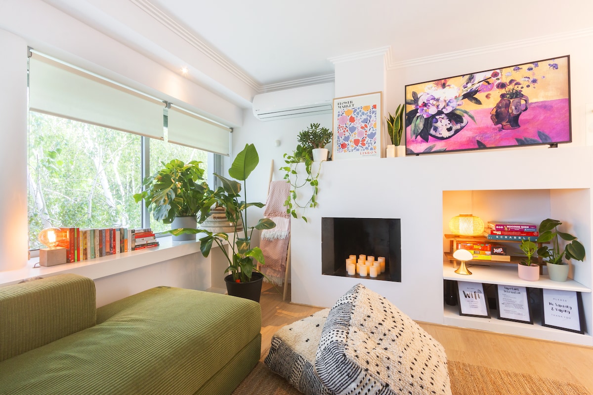 A cozy living area features a green sofa alongside a large window, allowing natural light. Houseplants add a touch of greenery, while a bookshelf displays various books. A modern fireplace is set against the wall, complemented by decorative candles and colorful wall art.