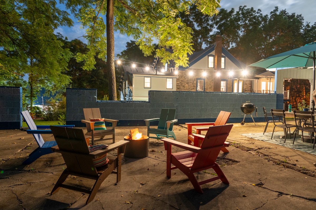 A backyard area is highlighted by Adirondack chairs in various colors arranged around a fire pit. String lights are overhead, gently illuminating the space. The house with a stone exterior and large windows is visible in the background, creating a welcoming evening atmosphere.