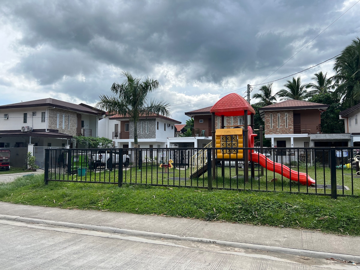 A fenced playground is seen across the street, featuring a colorful play structure with slides and climbing areas. Lush green grass surrounds the playground, with palm trees in the background and residential houses visible on either side.