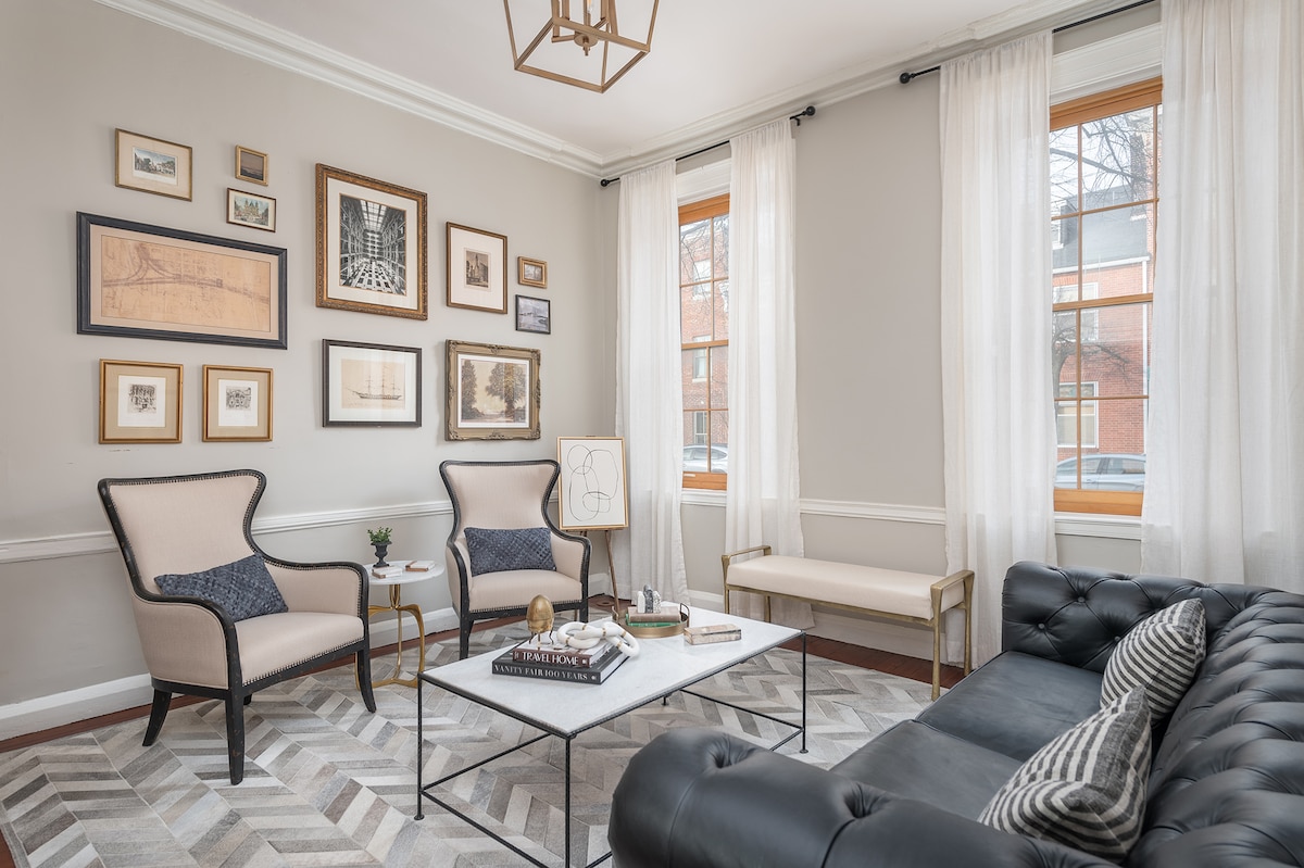 An elegantly designed sitting room features a vintage leather Chesterfield sofa and two accent chairs, complemented by a gallery wall of framed artworks. Natural light filters through sheer white curtains, illuminating a coffee table with decorative items, while a light-colored bench offers additional seating.