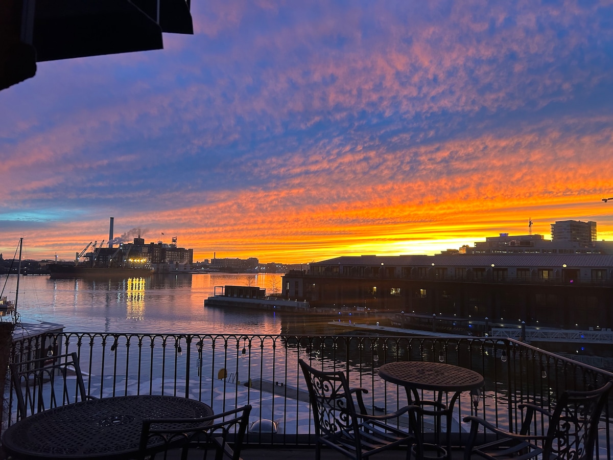 A breathtaking sunset illuminates the sky in vibrant shades of orange, pink, and purple over a calm harbor. A wrought iron balcony with a small round table and chairs frames the scenic view, adding an intimate touch to the picturesque waterfront scene.
