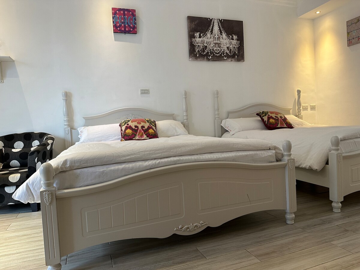 The room features two twin beds with light bedding arranged against a neutral wall. Colorful decorative pillows are placed on each bed, while a small, patterned chair is situated in one corner, adding a touch of contrast to the overall decor.