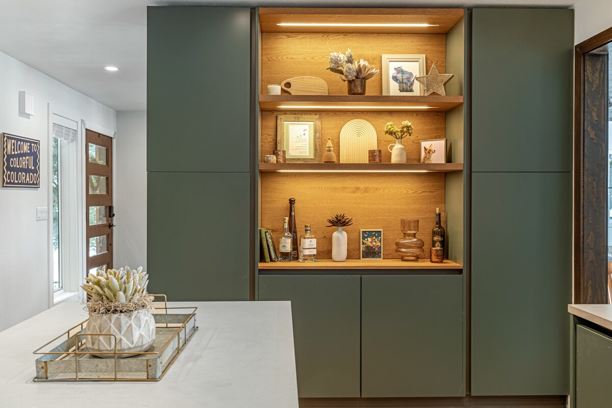 A modern kitchen area is highlighted by a well-organized display shelf featuring decorative items, plants, and framed photos. The cabinetry features a rich green color, complemented by warm wooden tones. A spacious countertop with subtle decor provides a functional and stylish workspace.