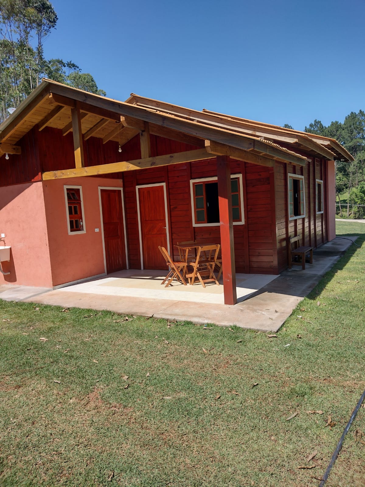 An exterior view of a rustic chalet features warm wooden accents and a covered porch. Two wooden doors are visible, alongside a small table with two chairs. A spacious grassy area surrounds the chalet, framed by tall trees in the background.