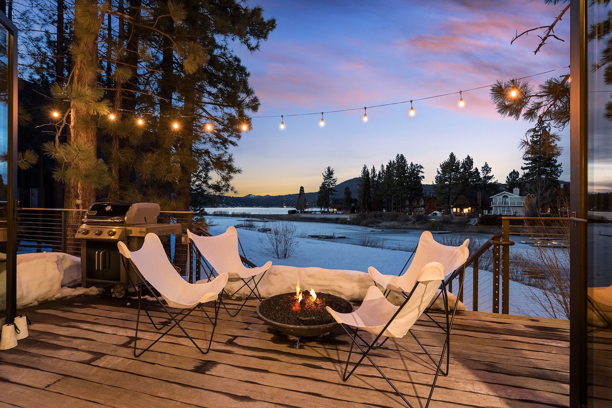 A spacious outdoor deck is equipped with comfortable seating arranged around a fire pit. A grill is visible nearby, and string lights create a warm ambiance against a backdrop of a serene lakeside view and towering trees.