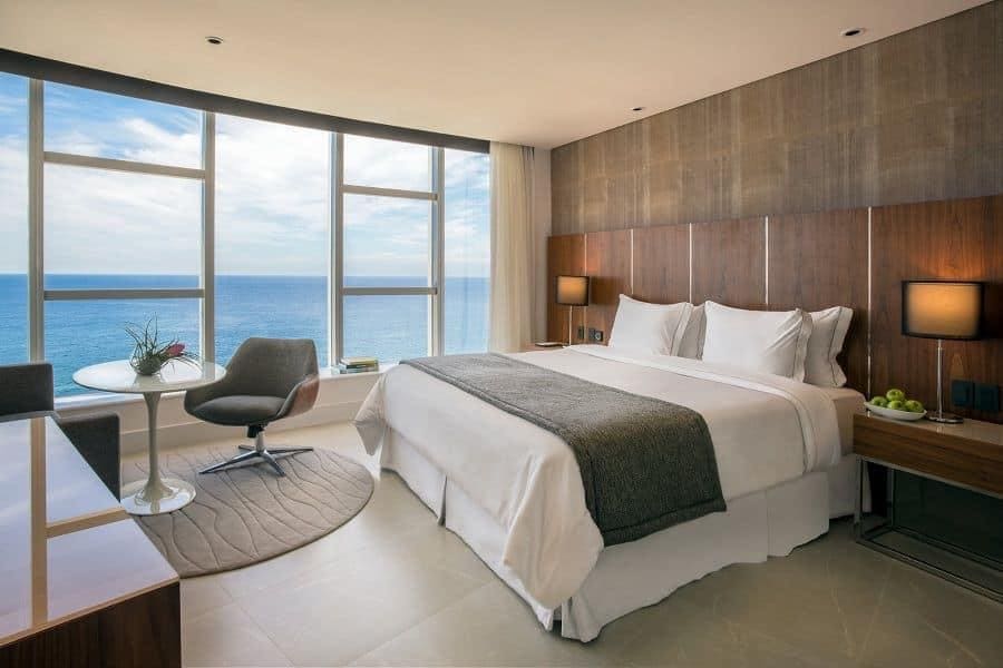 A spacious bedroom features a large bed with crisp white linens, complemented by wooden accents. A round table with a modern chair is positioned next to a large window offering a view of the ocean. Soft natural light fills the space.