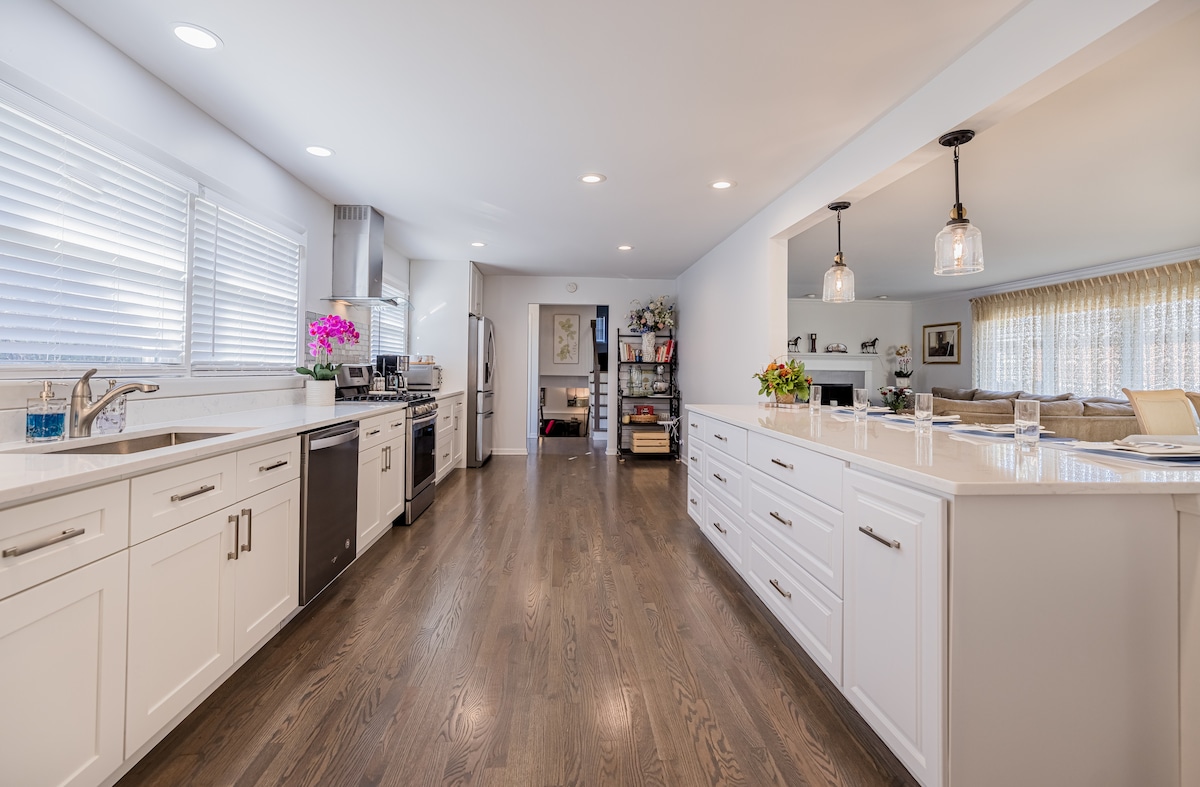 The kitchen features modern white cabinetry and sleek new appliances. A long island provides additional counter space, while natural light brightens the room through multiple windows. Adjacent to the kitchen, an open layout connects to a spacious dining and living area.