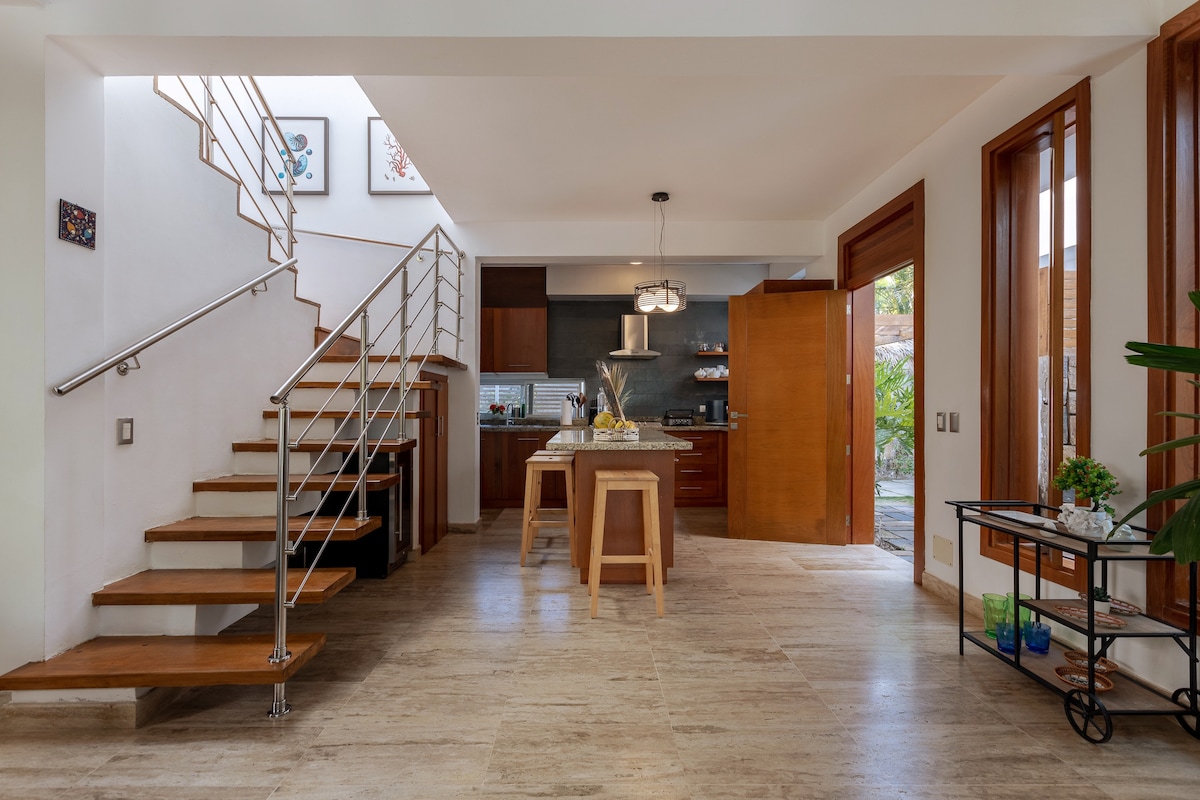 A bright interior space features a staircase with wooden steps and a metal railing. The open-plan layout connects a dining area with a kitchen seen in the background. Natural light filters through large windows, highlighting the neutral tones of the tiled floor.